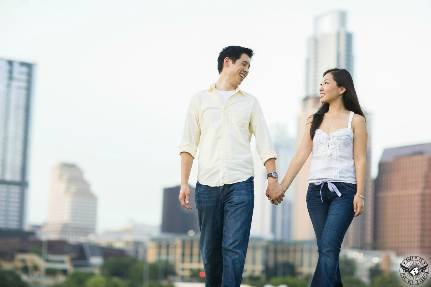 Petite Asian girl with long black hair wearing a white lacy tank top and hip hugger blue jeans holds hands with an Asian guy with dark spiky hair wearing a light yellow button up shirt with the sleeves rolled up and loose blue jeans walk near the Long Center the Austin high rise building in the background of this delightful engagement picture in Austin.